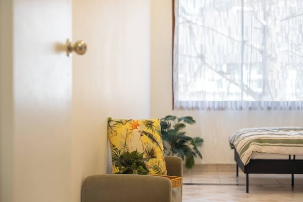 A bedroom with a bed, a chair with a tropical print cushion, a potted plant, and a window with a view of trees.