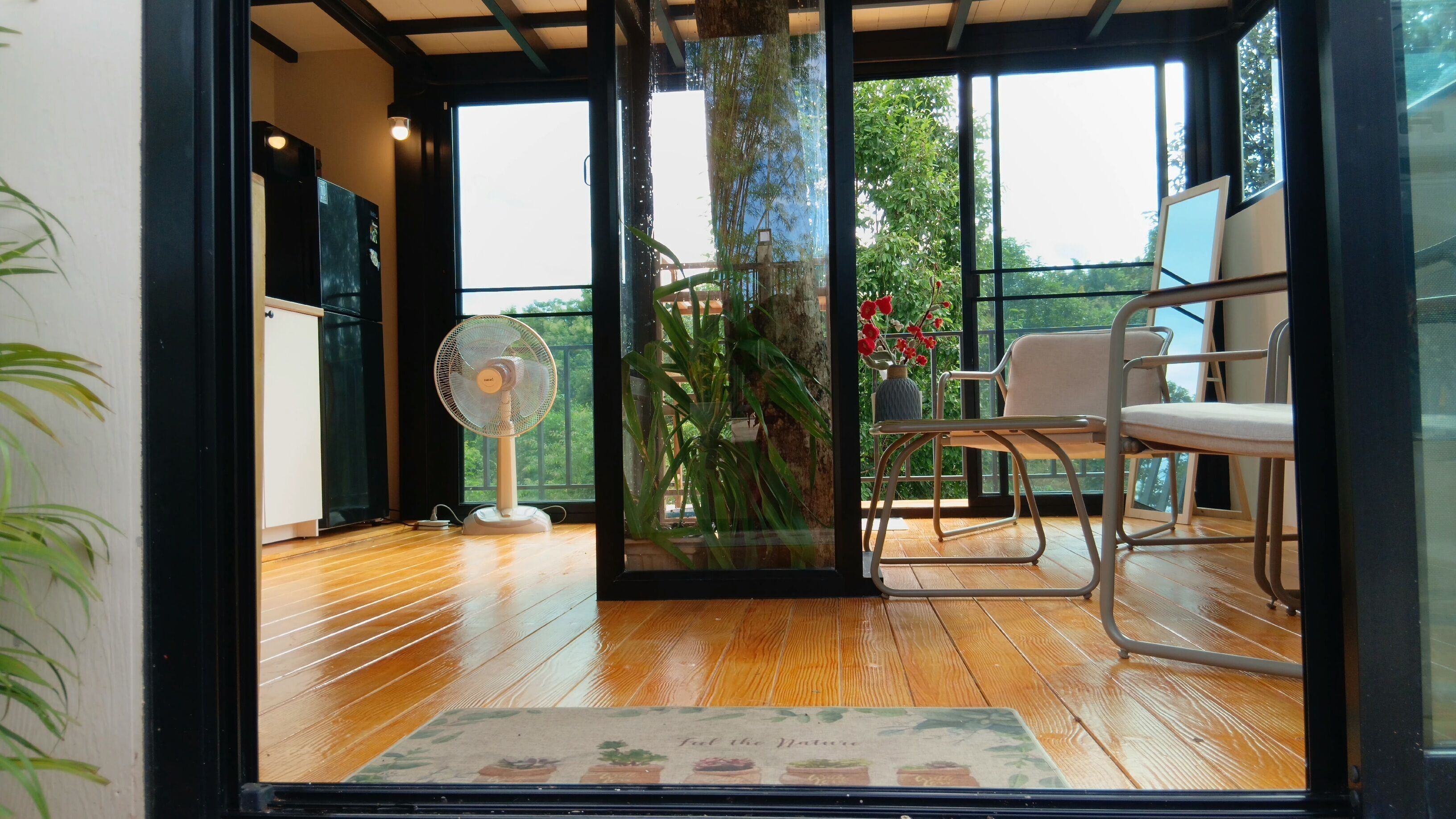 A room with a wooden floor, a standing fan, a table with chairs, and a view through a glass door to an outdoor area with plants and a railing.