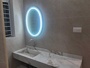 A bathroom with a marble sink, a modern faucet, and a wall-mounted oval mirror with a glowing light. - 7th Stay - Karaikudi (Karaikudi)
