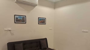 Een kamer met een donkerbruine bank, twee ingelijste foto’s aan de muur en een airconditioning.