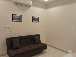 A room with a dark brown sofa, two framed pictures on the wall, and an air conditioning unit above. - 7th Stay - Karaikudi (Karaikudi)