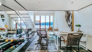 Un interior moderno con un escritorio de vidrio, dos sillas de mimbre, un ventanal con vistas y una escalera que lleva a un nivel superior.