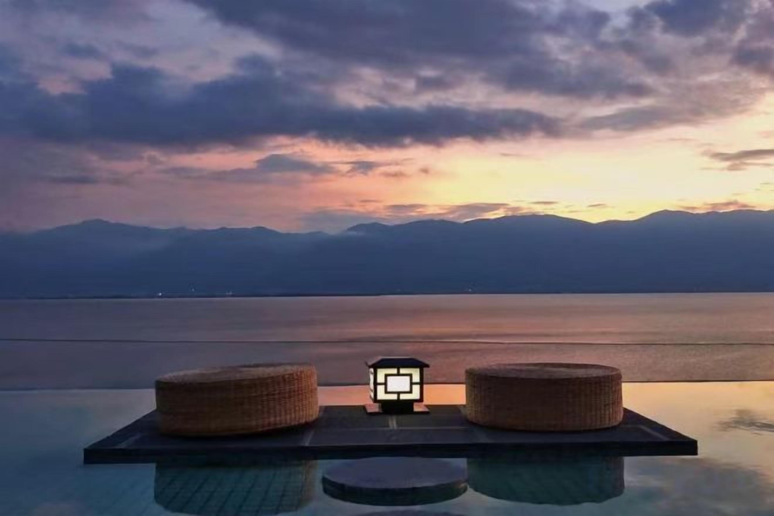 An infinity pool with two round wicker stools and a small illuminated object on a floating platform.