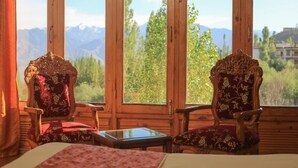 A room with two ornate chairs, a small table, and a bed. Large windows offer a view of mountains and trees. - The Druk Ladakh (Leh)