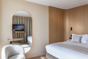 A modern hotel room with a bed, a TV mounted on a wooden panel, a round mirror, and a small seating area.