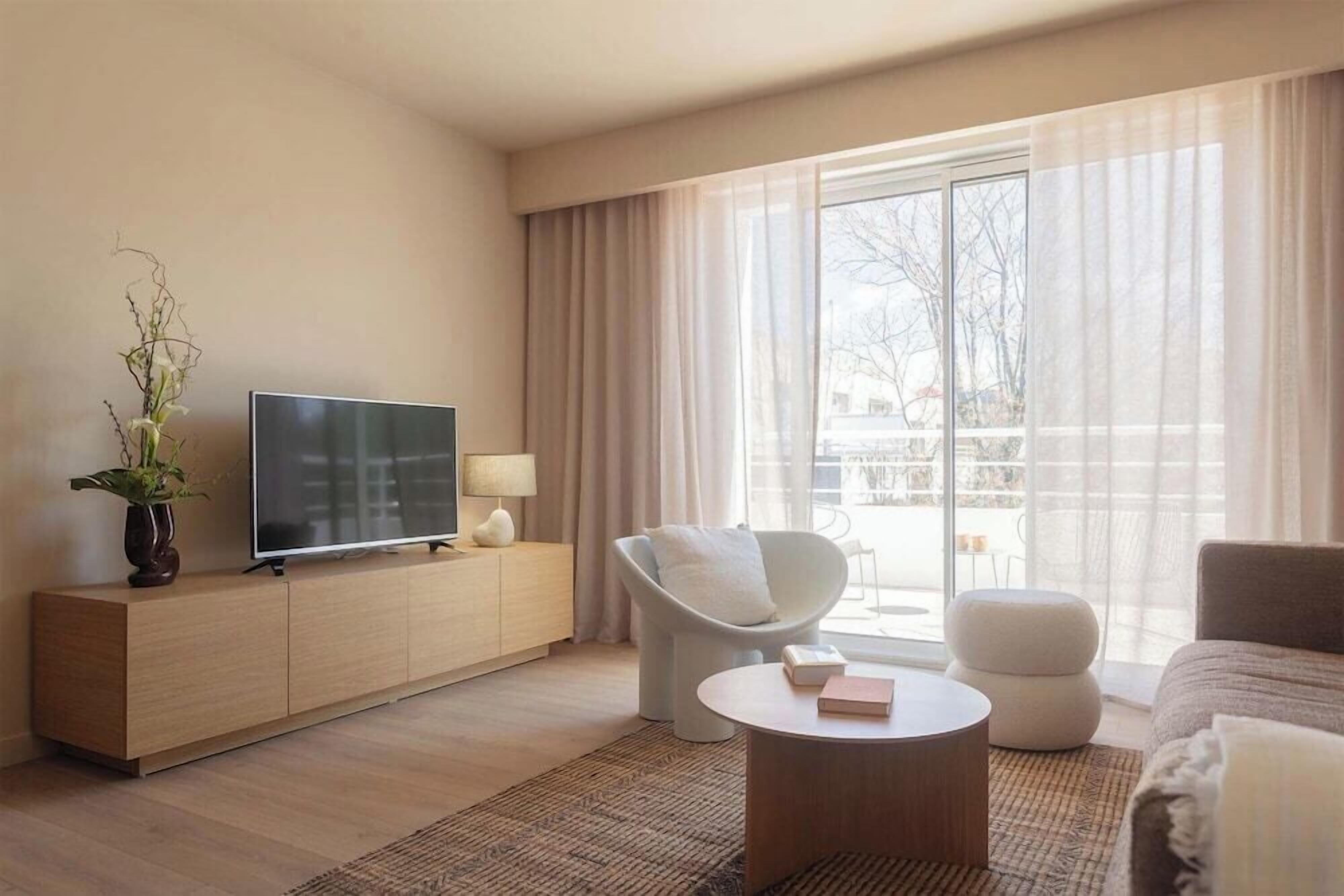 A modern living room with a flat-screen TV, a sofa, a chair, a coffee table, and a large window with curtains.