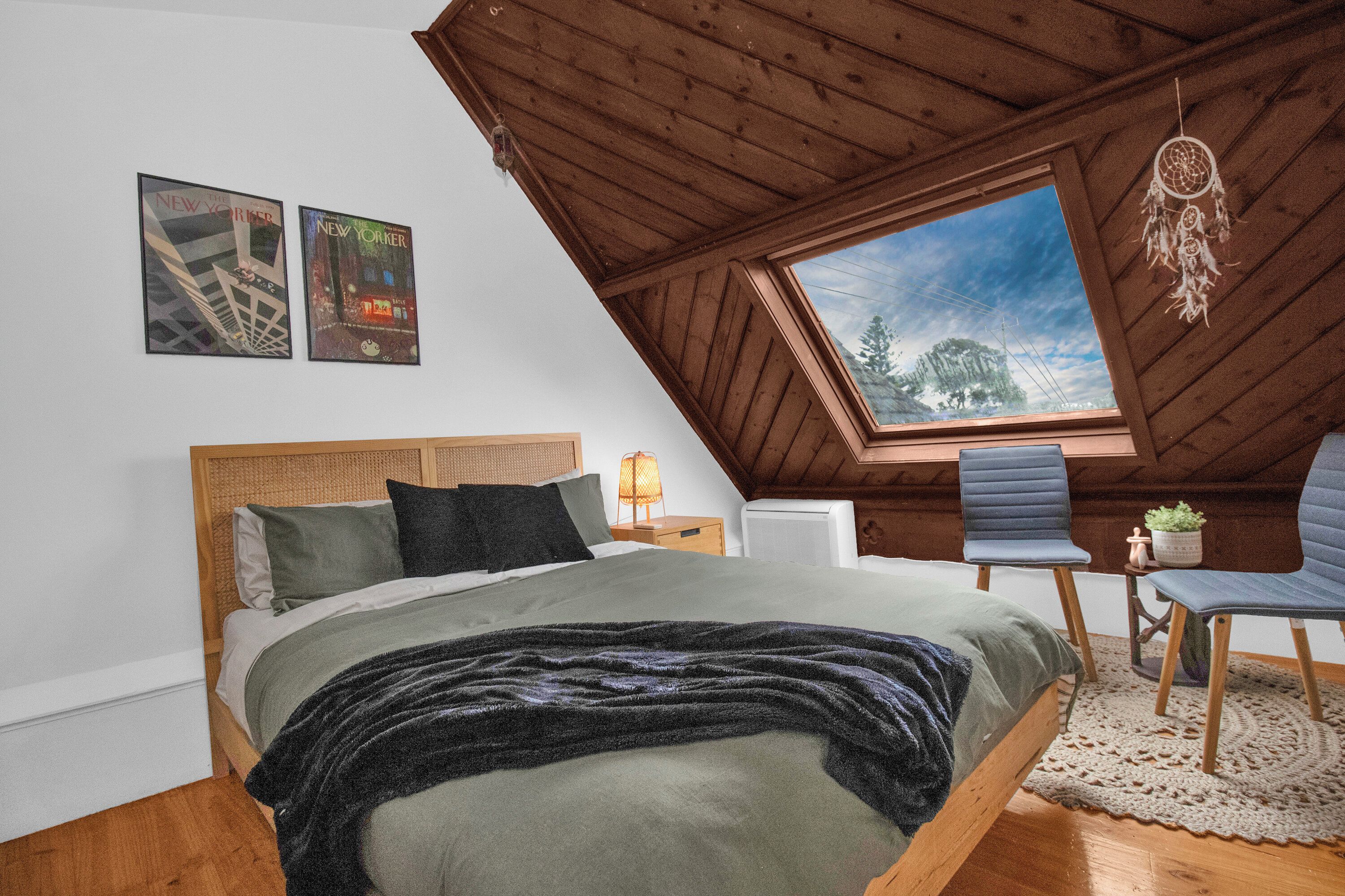 Une chambre avec un plafond en pente, un lit avec des oreillers, une chaise, un attrape-rêves et une fenêtre donnant sur un beau paysage.