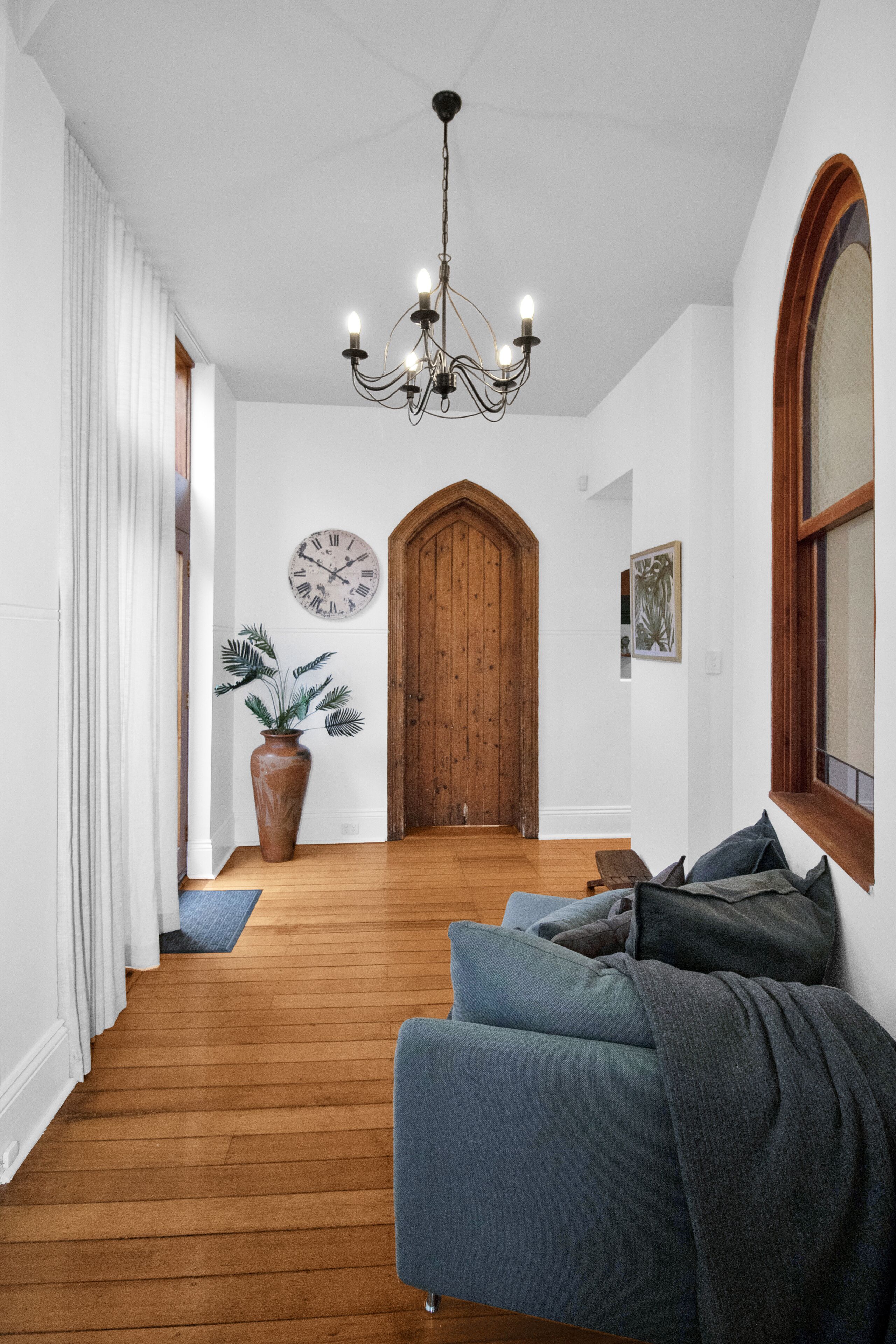 Un couloir avec un plancher en bois, un canapé bleu, une plante en pot, une porte en bois et une horloge murale.