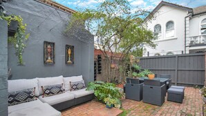 Eine Terrasse mit einer grauen Wand, einem weißen Sofa mit gemusterten Kissen, Korbmöbeln und Blumentöpfen.