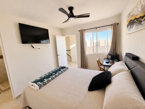 Ein Schlafzimmer mit einem Bett, einem Schreibtisch mit Computer, einem Deckenventilator, einem an der Wand montierten Fernseher und einem Fenster mit Vorhängen.