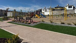 Ein Spielplatz mit Rutschen und Schaukeln, umgeben von einem gepflasterten Weg und Grünflächen.