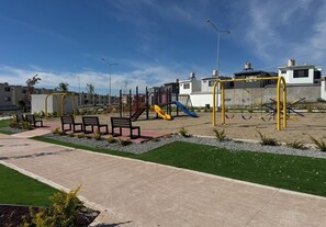 Ein Spielplatz mit Rutschen und Schaukeln, umgeben von einem gepflasterten Weg und Grünflächen.