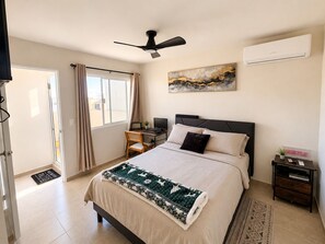 Habitación de hotel con cama, escritorio, silla, ventilador de techo, equipo de aire acondicionado y ventana con cortinas.