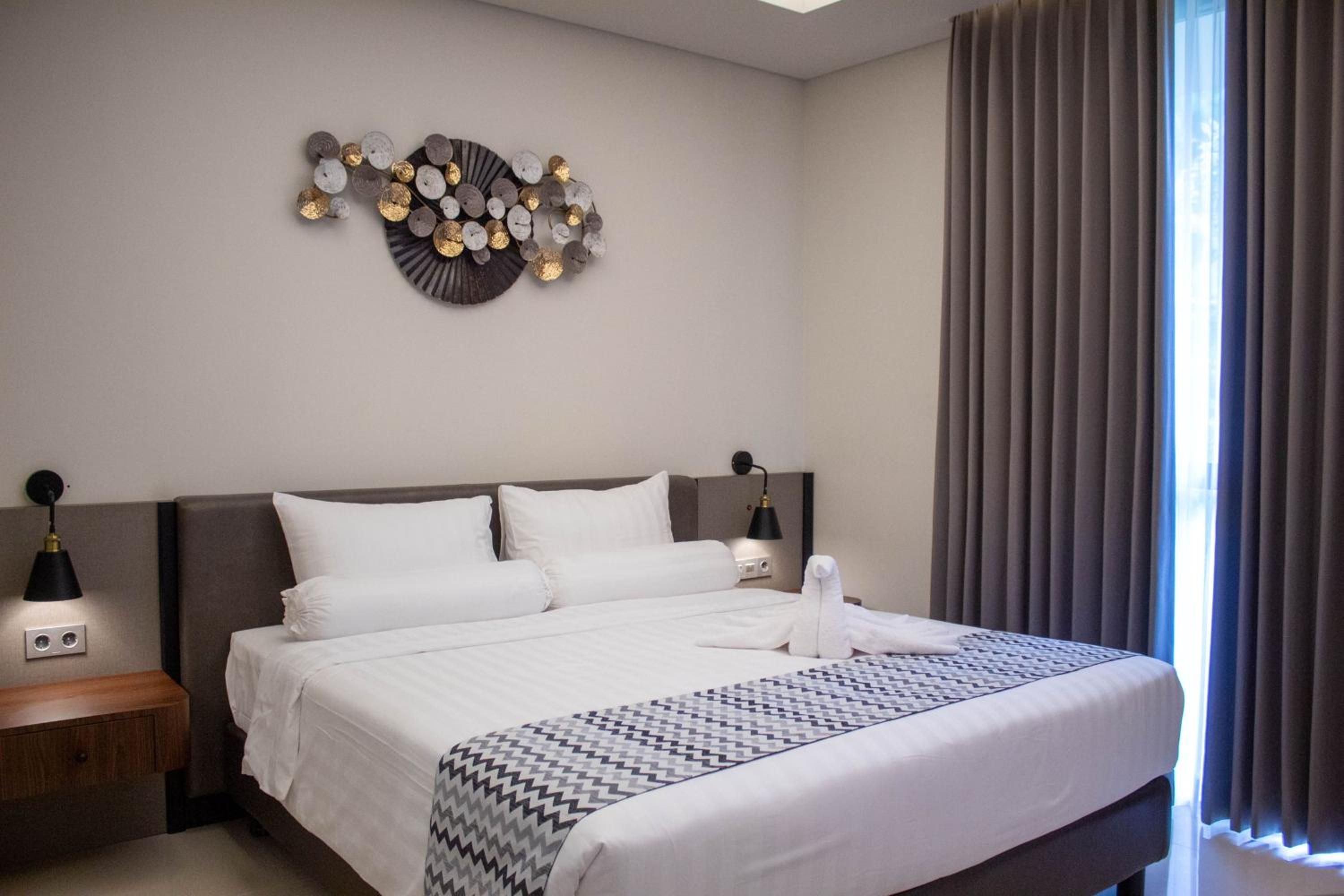 Ein Hotelzimmer mit einem großen Bett, zwei Nachttischen, einer Wandleuchte und einem fensterbepflanzten Fenster mit Vorhängen.