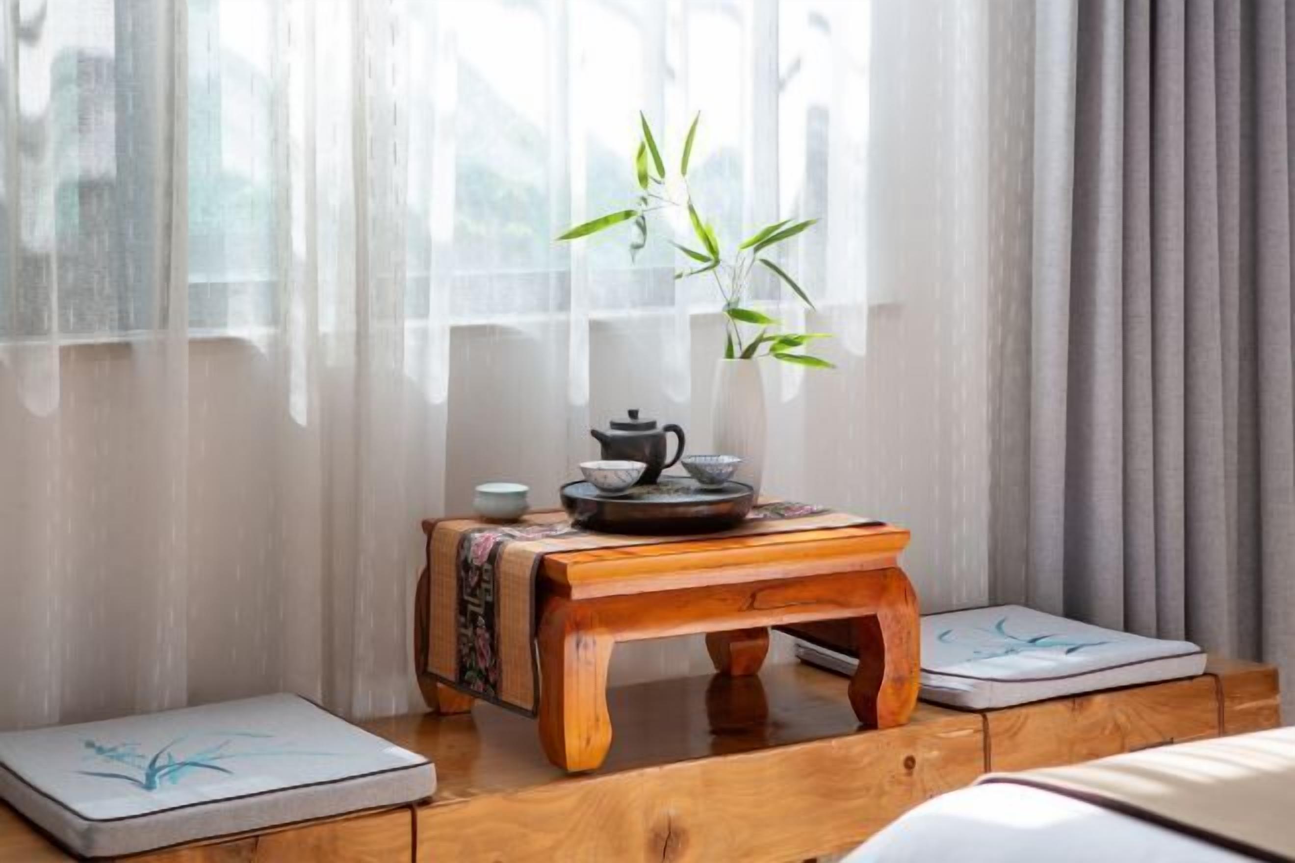 A room with a traditional tea set on a wooden table, a bamboo plant, and a window with sheer curtains.