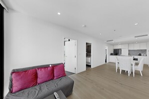 Ein modernes Wohnzimmer mit grauem Sofa, einem Essbereich mit weißen Stühlen und einer Küche mit weißen Schränken.