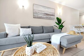 A modern living room with a grey sofa, a round wicker coffee table, and a painting on the wall. - Poolside Holiday Haven at Whitfield Waters (Manoora)