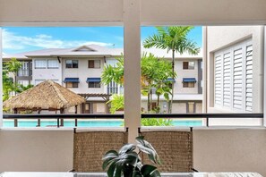 Ein Balkon mit Blick auf einen Swimmingpool, Palmen und ein Gebäude mit einem Reetdach.