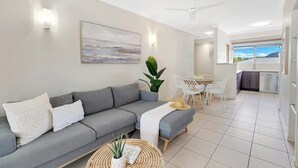 Uma sala de estar moderna com um sofá cinzento, uma área de refeições com uma mesa e cadeiras brancas, e uma cozinha ao fundo.