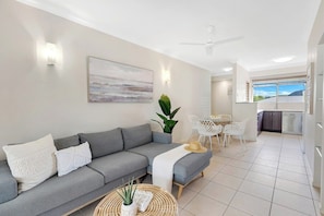 Ein modernes Wohnzimmer mit einem grauen Sofa, einem Essbereich mit einem weißen Tisch und Stühlen sowie einer Küche im Hintergrund.