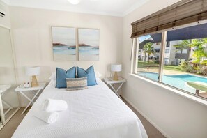 Ein Hotelzimmer mit einem Bett, zwei Nachttischen, einem Fenster mit Blick auf einen Swimmingpool und Gebäude sowie zwei an der Wand befestigten Bildern.
