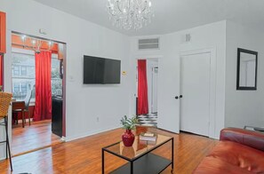 A modern living room with a flat-screen TV, a glass coffee table, a red sofa, and a chandelier. - 1BR The Montrose W Main 02, Downtown, Med Center (Houston)