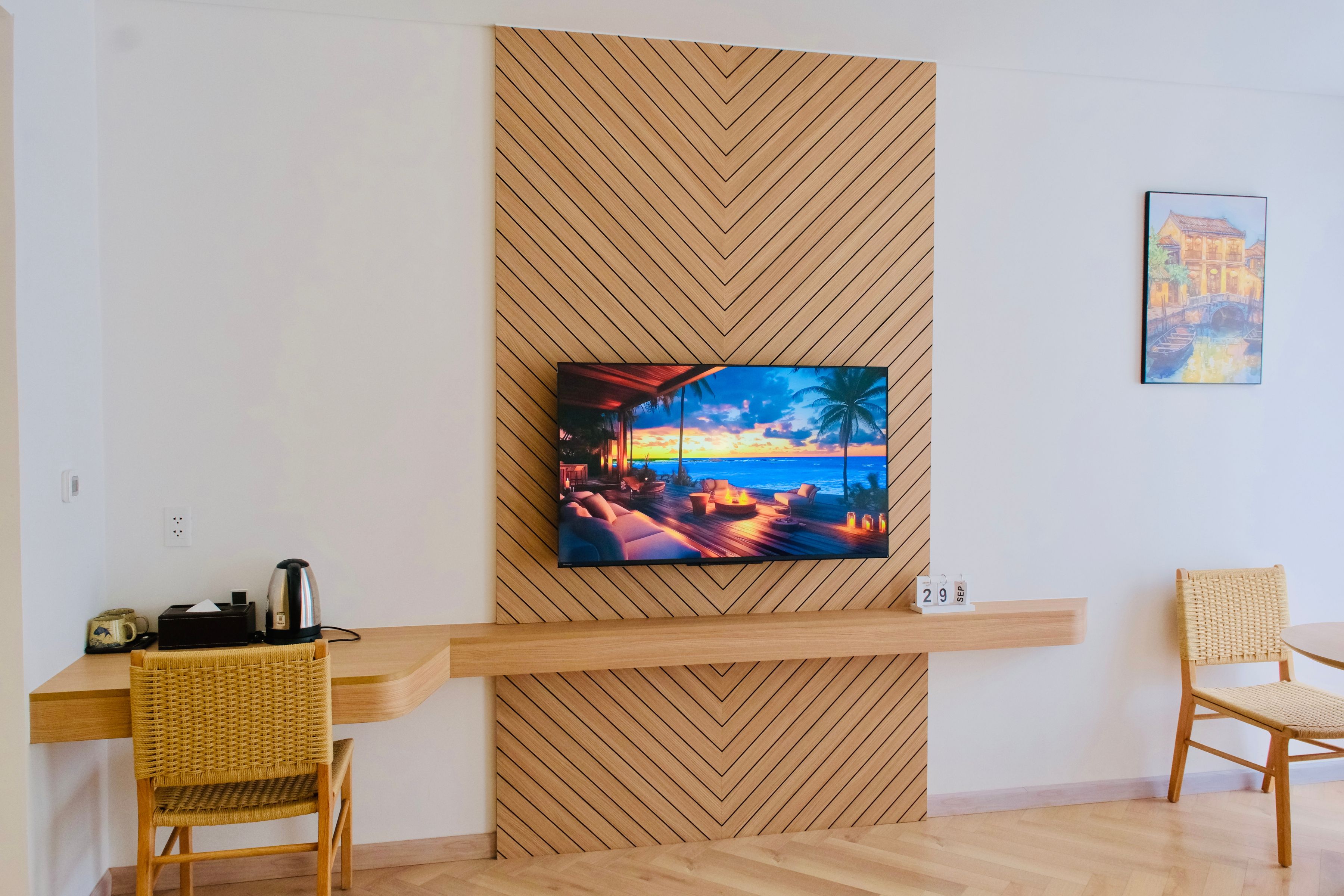 A modern room with a wooden herringbone wall, a flat-screen TV mounted on it, a wooden desk, two wicker chairs, and a small shelf with a date calendar.