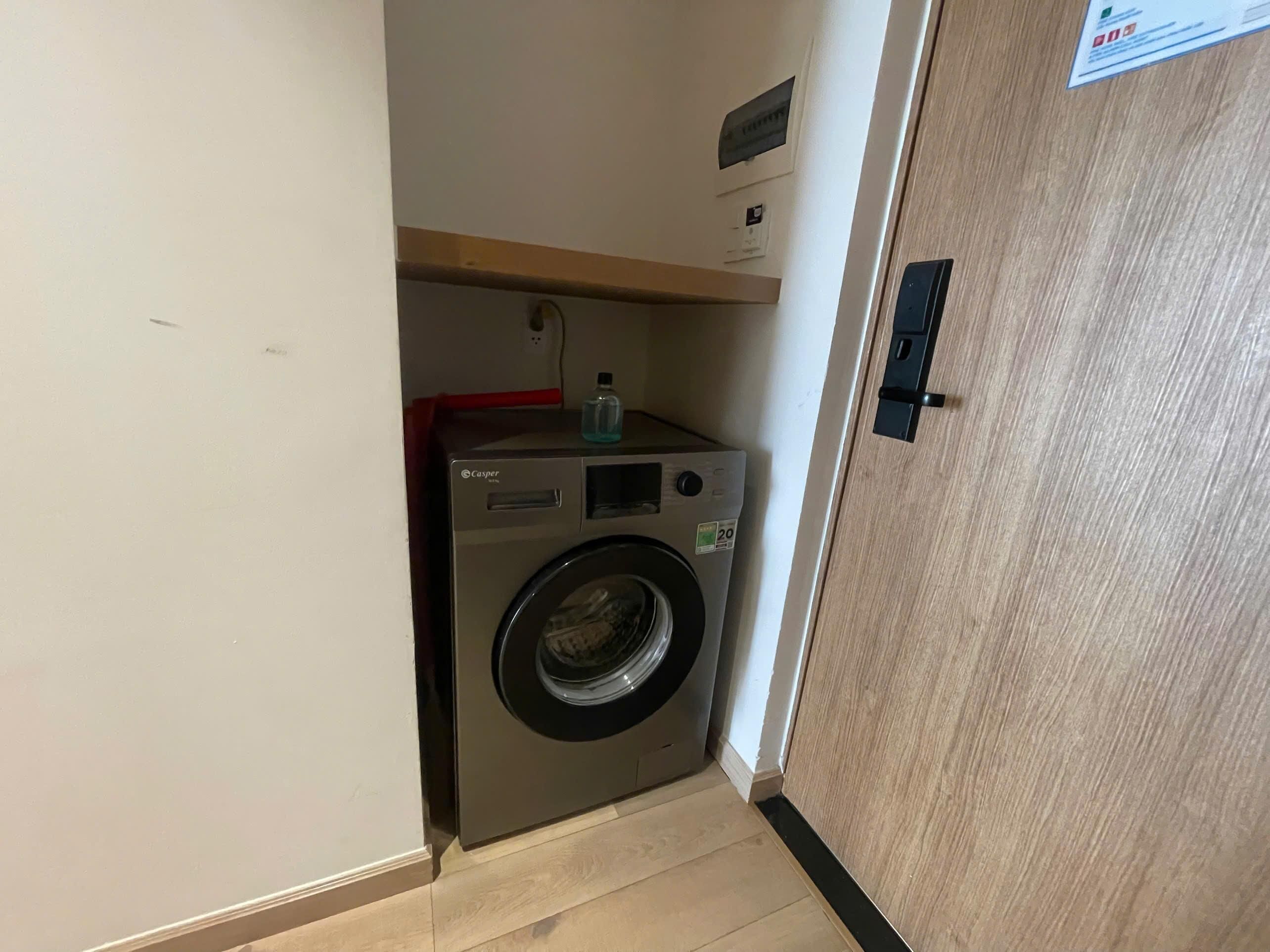 Une petite buanderie moderne, dotée d’un lave-linge à chargement frontal, d’une petite étagère avec une bouteille et d’une porte en bois.