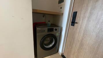 Une petite buanderie moderne, dotée d’un lave-linge à chargement frontal, d’une petite étagère avec une bouteille et d’une porte en bois.