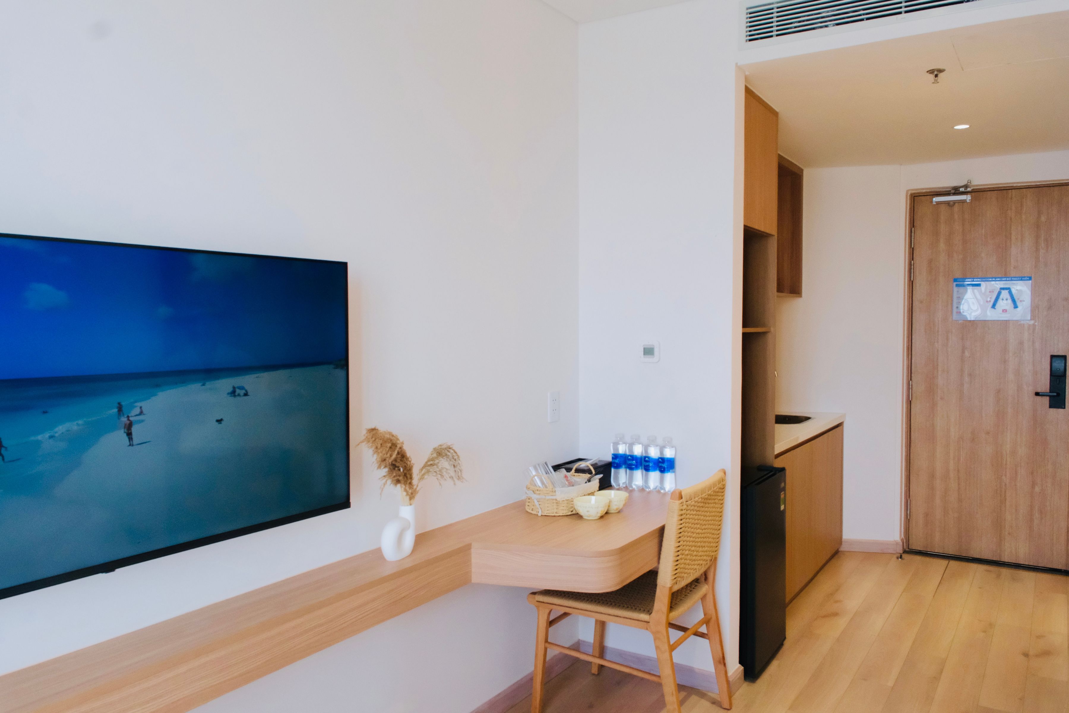 A room with a large flat-screen TV on the wall, a wooden table with a chair, a basket, and bottled water.