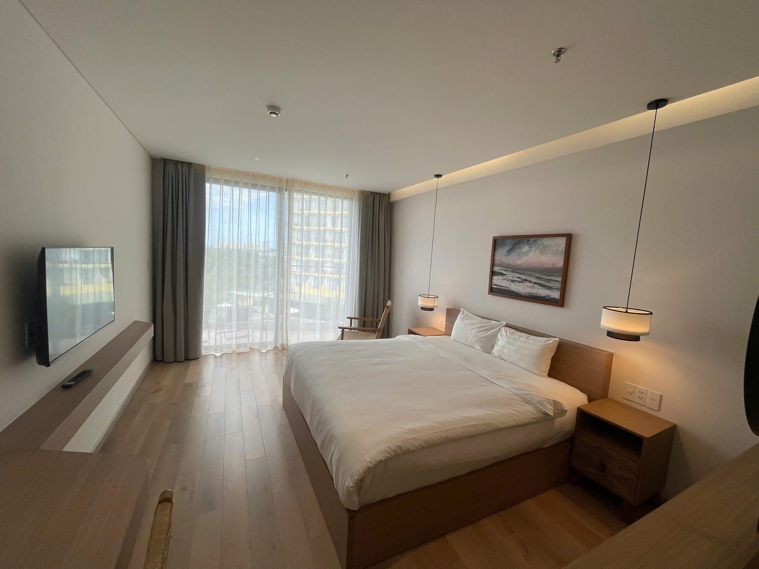 A modern hotel room with a large bed, a flat-screen TV, a painting, and a balcony with curtains.