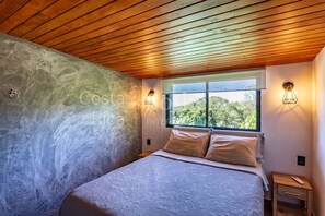 Ein Schlafzimmer mit Bett, Nachttisch, Fenster mit Blick und Holzbalkendecke.
