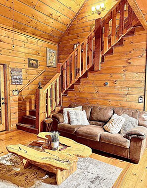 A log cabin interior with a wooden staircase, a brown leather sofa, and a coffee table. - Charming Cabin in the clouds with hot tub, screen porch and fireplace (Ellijay)