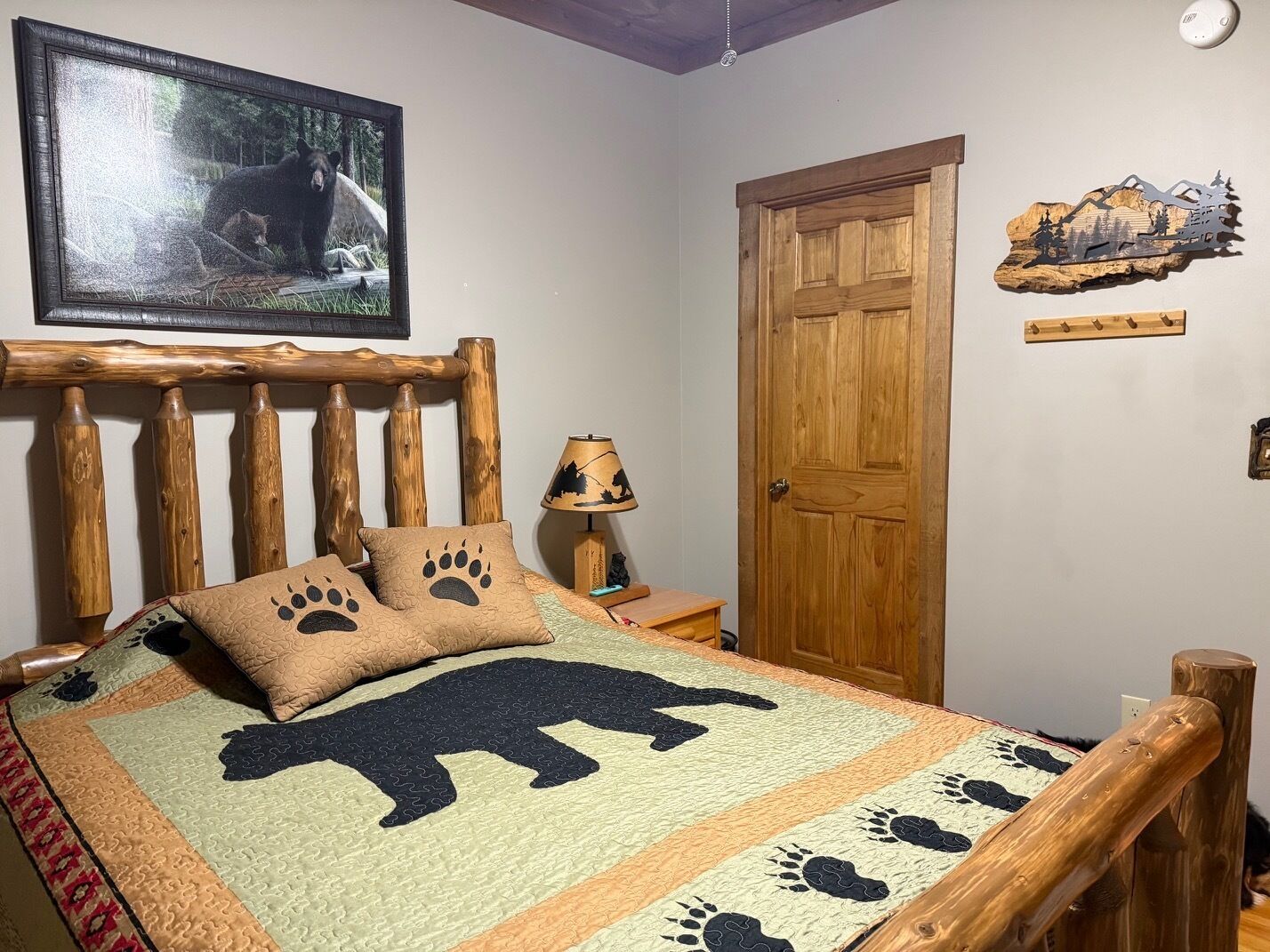 A bedroom with a wooden headboard, a bed with a bear-themed quilt, a nightstand with a lamp, and a door with a wooden panel.