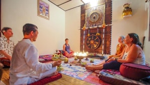 A group of people seated on a patterned rug in a room with a large decorative wheel on the wall.