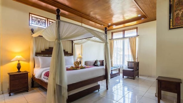 A four-poster bed with a canopy, a nightstand with a lamp, a wooden bench, and a small table in a well-lit room with tiled flooring and a wooden ceiling.