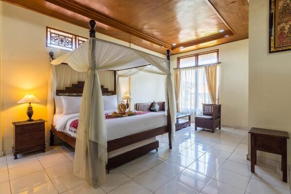 A four-poster bed with a canopy, a nightstand with a lamp, a wooden bench, and a small table in a well-lit room with tiled flooring and a wooden ceiling.