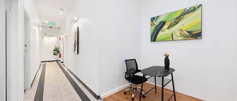 A hallway with a tiled floor, a black office chair, a small table, and a painting on the wall.