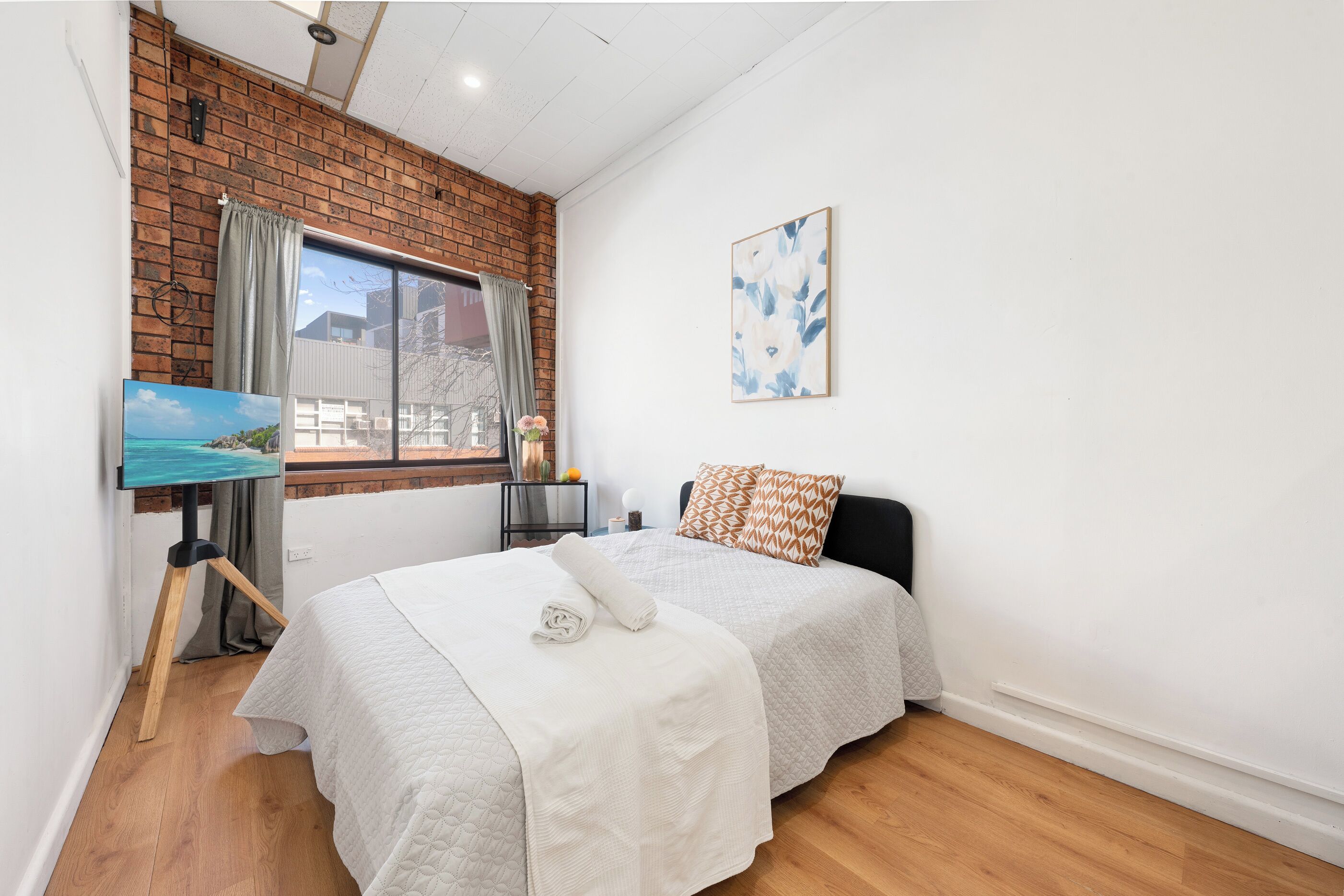 A bedroom with a bed, a painting, a window with curtains, and a wall-mounted picture.