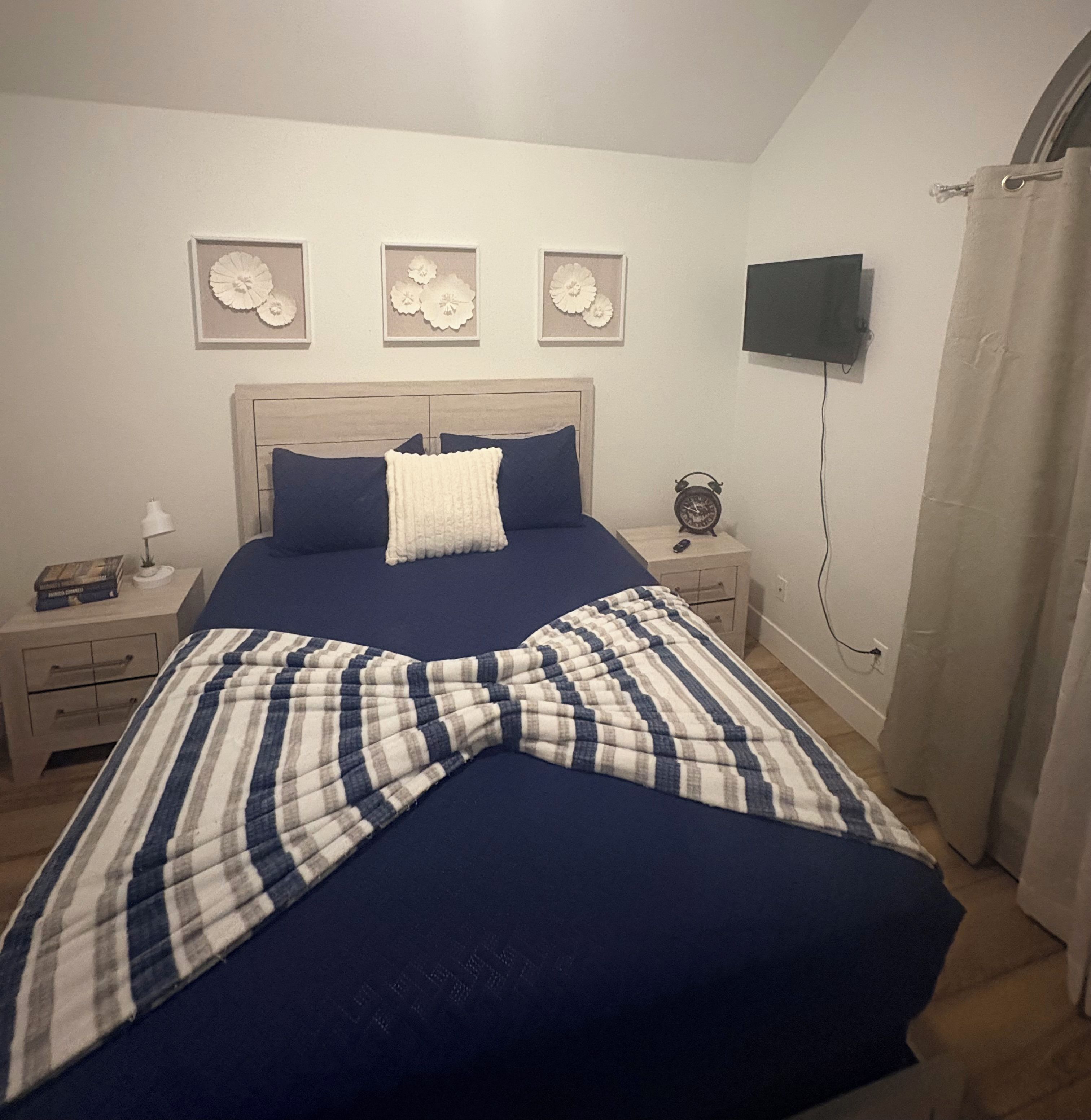 A bedroom with a bed, a bedside table, a lamp, a clock, and a TV mounted on the wall.