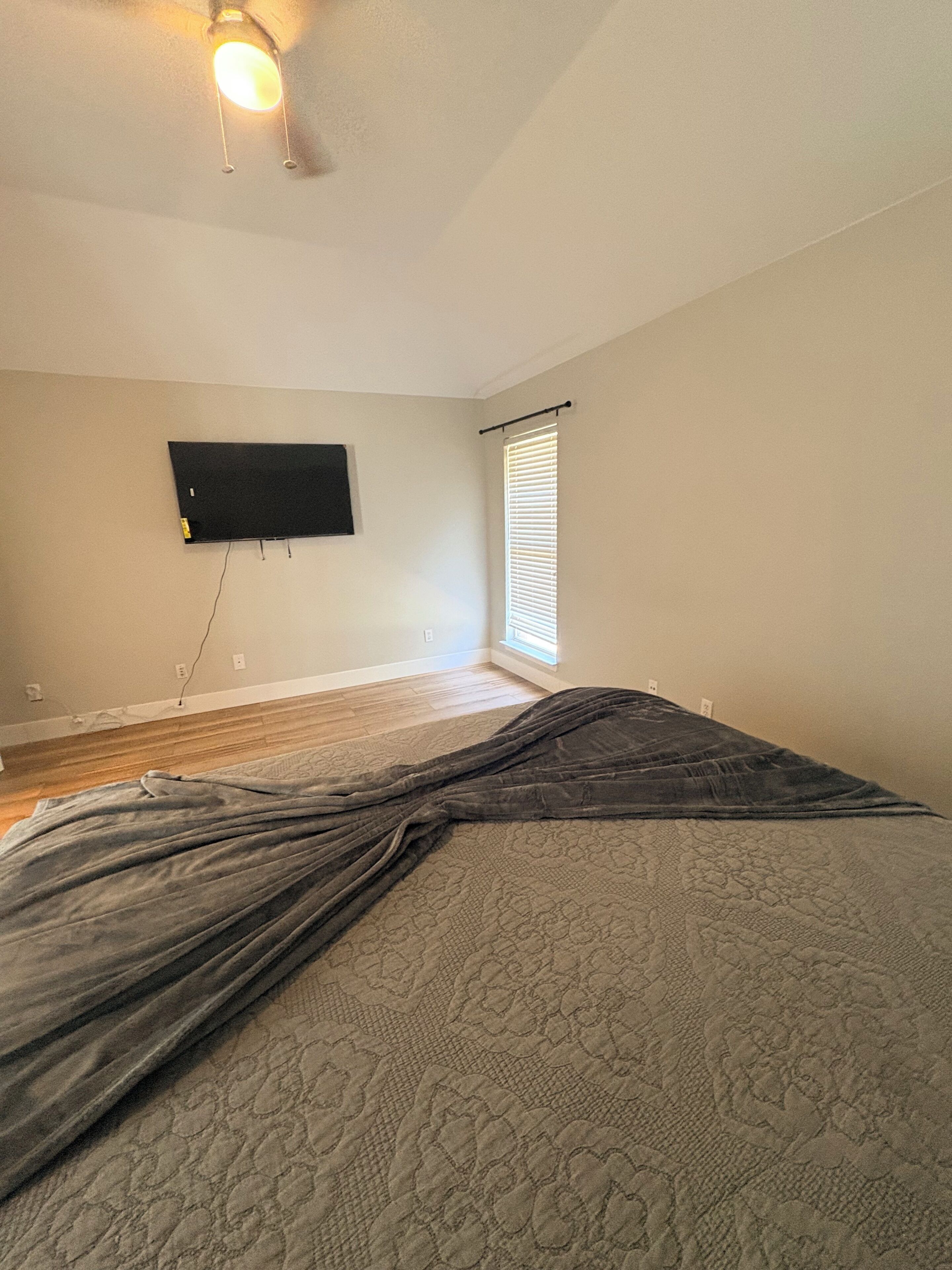 A room with a bed, a television mounted on the wall, and a window with blinds.