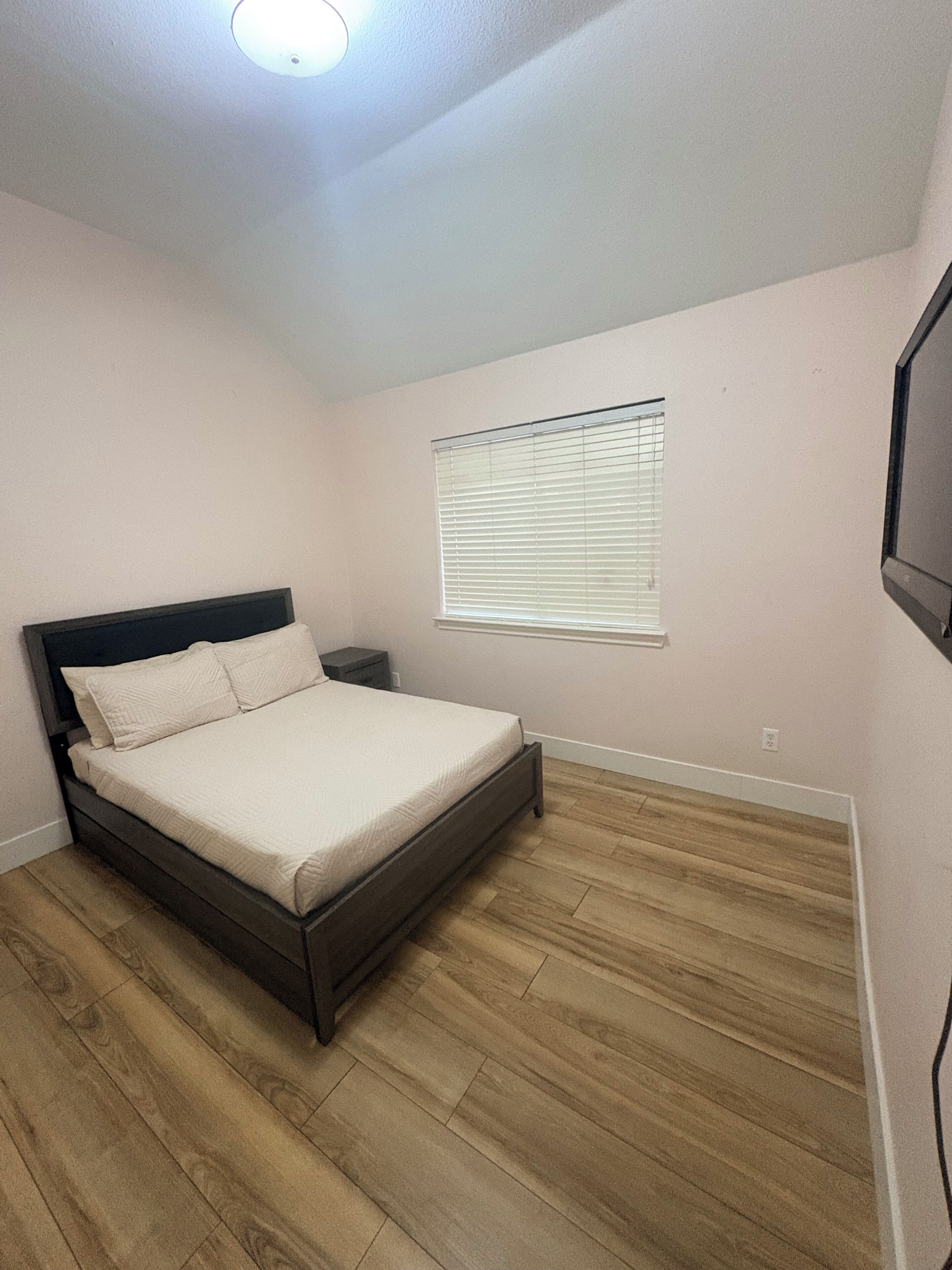 A bedroom with a bed, a nightstand, a window with blinds, and a ceiling light.