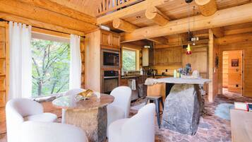 A cabin interior with a stone table, white chairs, a kitchen area, and a view of trees through the window.