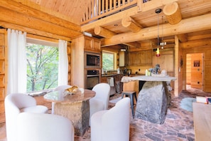 A cabin interior with a stone table, white chairs, a kitchen area, and a view of trees through the window.