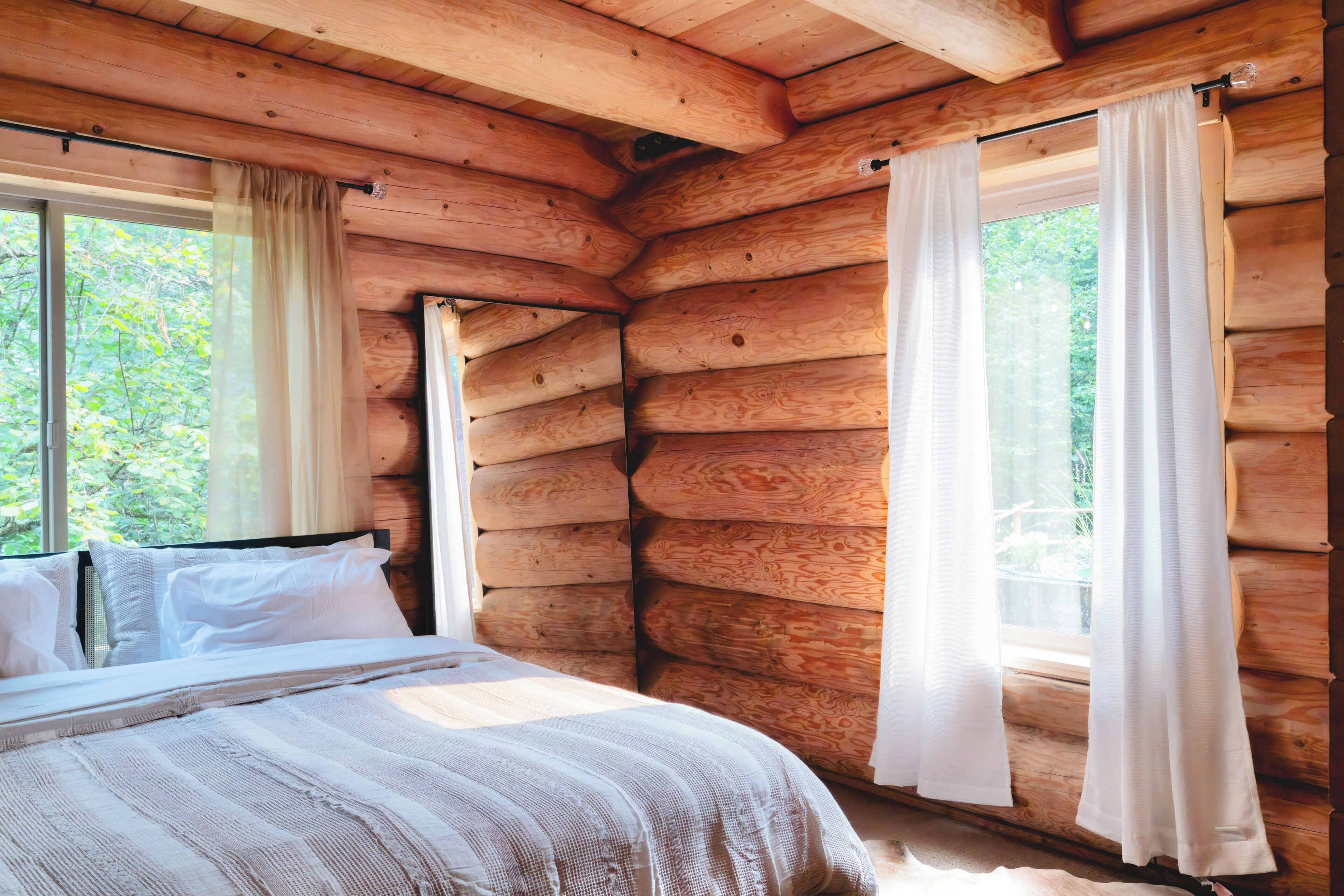 A log cabin bedroom with a bed, curtains, and a window overlooking greenery.