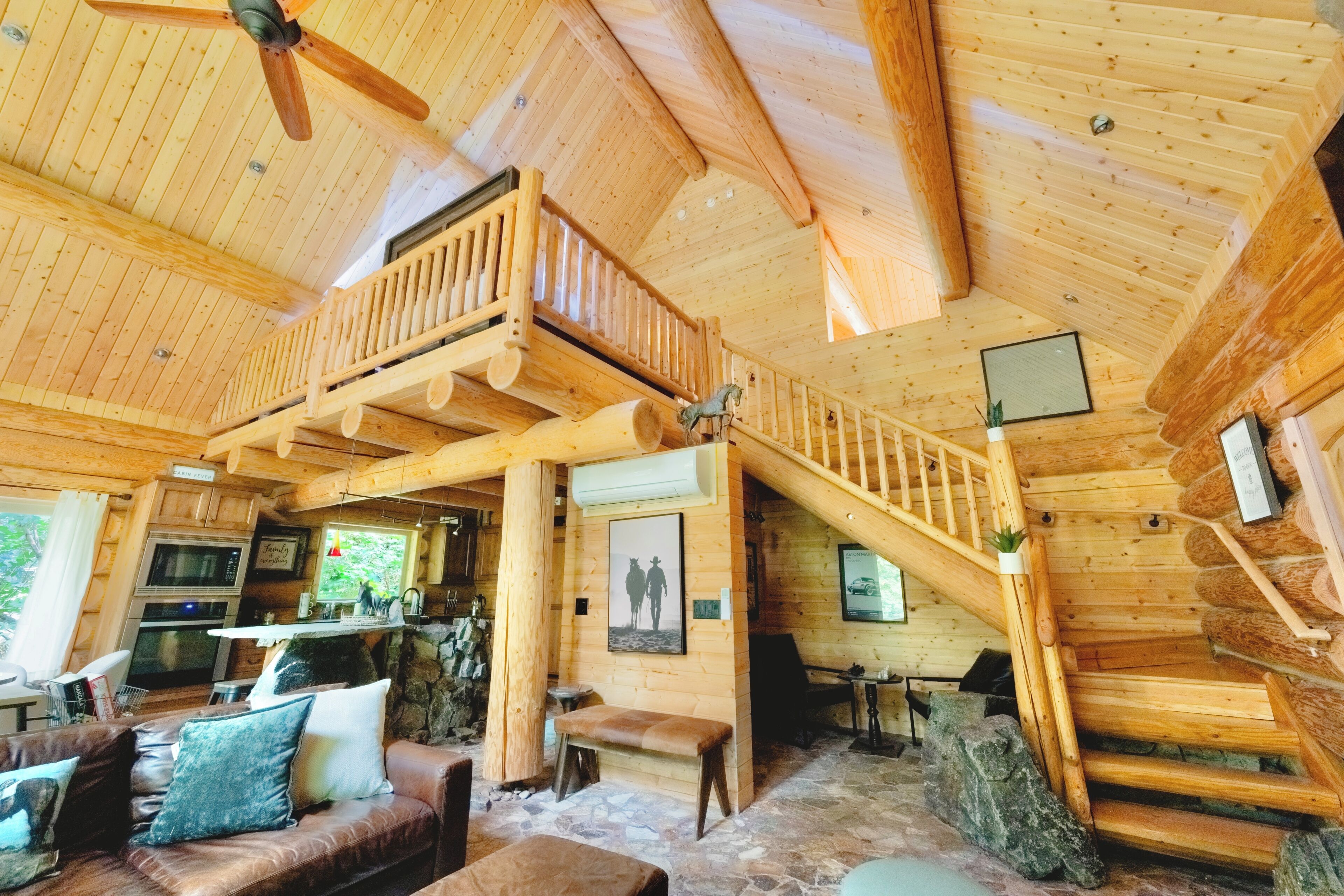 A cabin interior with a wooden staircase, a balcony, a fireplace, and a stone fireplace surround.