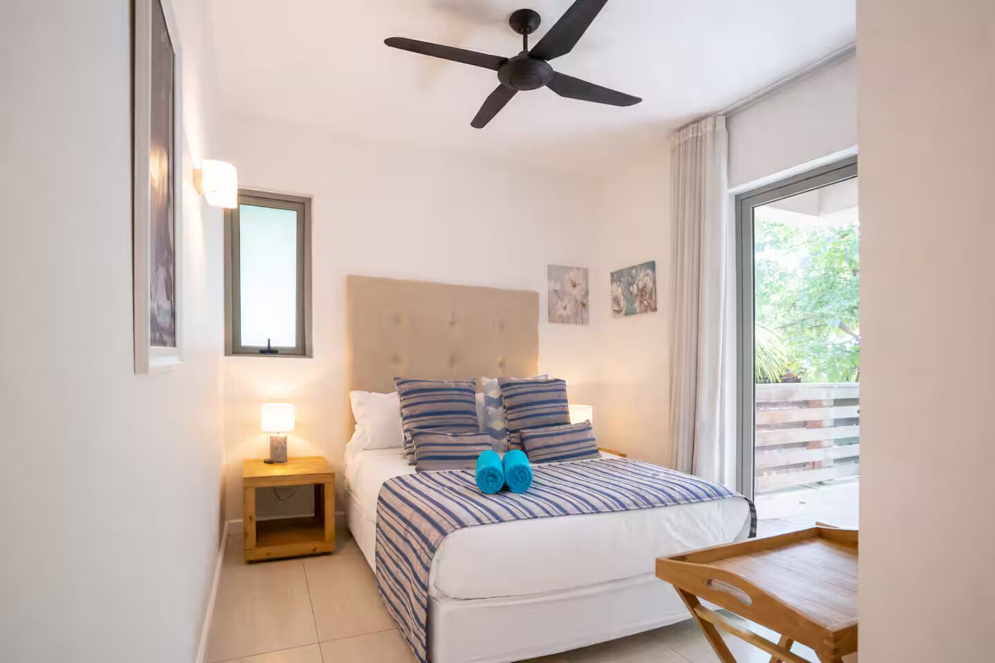 Quarto com cama, mesa de cabeceira, ventilador de teto, janela e vista para o verde lá fora.