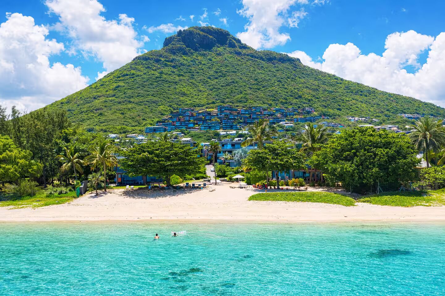 Un complexe hôtelier de bord de mer, avec des montagnes en toile de fond, une eau turquoise cristalline et une plage de sable.