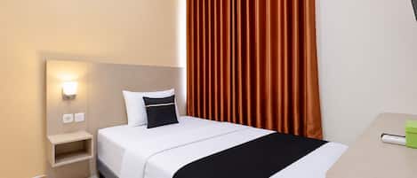 A hotel room with a bed, a nightstand with a lamp, a desk with a green container, and a window with brown curtains.