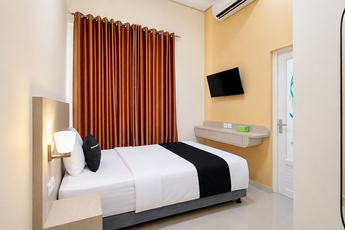 A hotel room with a bed, a flat-screen TV, an air conditioning unit, and a bedside table with a lamp.